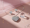 Heart-shaped silver necklace and earrings on a pink surface with scattered diamonds.